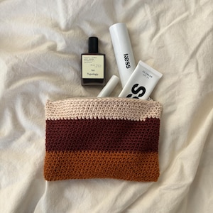 Könnte beinhalten: Gehäkelte Kosmetiktasche mit Reißverschluss und horizontalen Streifen in Beige, Burgunderrot und Orange. Die Tasche enthält Schönheitsprodukte, darunter eine Typology-Parfümflasche, Kess-Sonnencreme und eine CC-Creme-Tube.