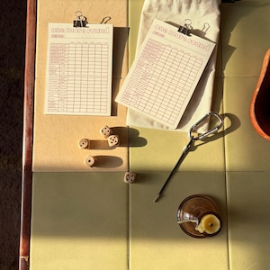 May include: Two cream-coloured scorecards with the text one more round and a grid, four small wooden dice, a beige drawstring pouch, and a silver carabiner with a thin metal rod attached, all arranged on a light green tiled surface.