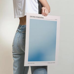 May include: A rectangular print with a soft blue gradient, labelled "DIVINE FEMININE" at the top and "AURA COLLECTION" at the bottom. The print is held by a person wearing a white top and blue jeans.