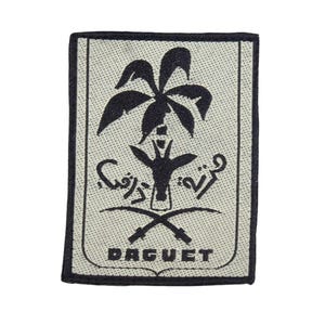 Pode incluir: Um remendo bordado em preto e branco com uma palmeira, espadas cruzadas e texto em árabe. O texto diz "BAGUET".