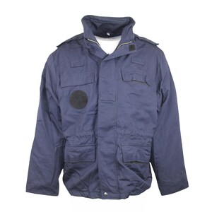 May include: Navy blue jacket with a high collar, multiple pockets, and a black circular patch on the left chest. The jacket has a zipper closure and epaulets on the shoulders. The jacket appears to be made of a durable, possibly water-resistant material.