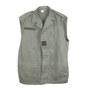 Op de afbeelding: Een mouwloos, olijfgroen vest met een kraag en knoopsluiting. Het vest heeft twee borstzakken met ritssluiting, twee onderste klepzakken en een kleine patch op de borst. Het kledingstuk hangt aan een hanger.