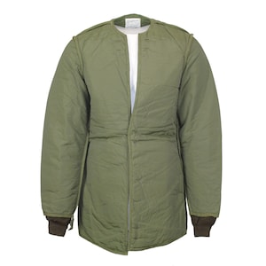 Op de afbeelding: Olijfgroene gewatteerde militaire jas met een ronde kraag en een lange ritssluiting. De jas heeft een gewatteerd ontwerp, bruine geribde manchetten en een wit ondershirt. Ontworpen om als laag te worden gedragen.