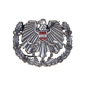 Peut inclure: Insigne métallique argenté représentant un aigle aux ailes déployées, un écusson à rayures rouges et blanches, et une couronne de feuilles de chêne. L'insigne est doté d'une chaîne et d'un fermoir à crochet.