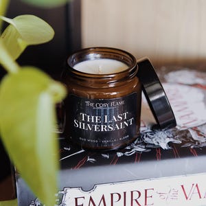 May include: A brown glass candle jar with a white candle inside. The jar has a black label that reads "The Cosy Flame" and "The Last Silversaint" with the scent notes "Oud Wood - Vanilla - Blood". The candle is sitting on top of a book with the title "Empire of the Va" printed on the cover.