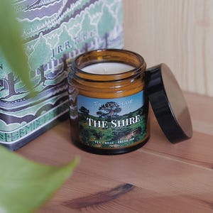 Könnte beinhalten: Ein braunes Glasgefäß mit einer Kerze und einem schwarzen Deckel. Das Etikett auf dem Gefäß lautet "The Cosy Flame" und "The Shire" mit dem Text "Tea - Rose - Fresh Air" darunter.