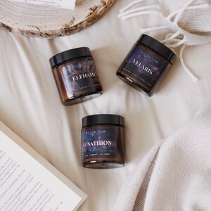 May include: Three amber glass candle jars with black lids. The labels are black with white text. The candles are labeled "Elfhame", "Velaris", and "Lunathion".