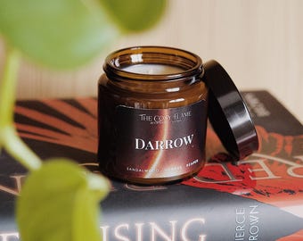 May include: A brown glass candle jar with a black label that reads "The Cosy Flame" and "Darrow" in white text. The candle is on top of a stack of books with the title "Raising Son" visible.