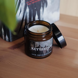 Puede incluir: Un tarro de vela de vidrio marrón con una etiqueta blanca que dice "The Cosy Flame" y "Ketterdam" en negro. La etiqueta también incluye el texto "Coffee - Tobacco Leaves - Crown".