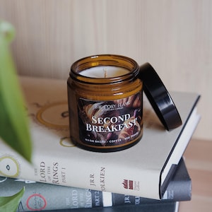 Può includere: Un barattolo di vetro marrone per candele con un coperchio nero. L'etichetta recita "The Cosy Flame" e "Second Breakfast" con il testo "Warm Bread, Coffee, Red Fruit" sotto. La candela è appoggiata su una pila di libri con il titolo "Il Signore degli Anelli" visibile.