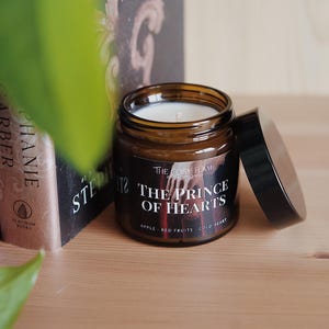 May include: A brown glass candle jar with a black lid. The label reads "The Cosy Flame" and "The Prince of Hearts" with the scent notes "Apple, Red Fruits, Cold Heart".