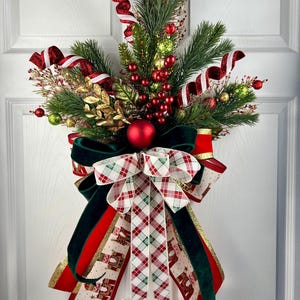 May include: A festive Christmas door swag featuring faux evergreen branches, red berries, and gold accents. The swag is adorned with a large bow made of red, green, and plaid ribbons. A red ornament is centered in the design, creating a holiday-themed decoration.