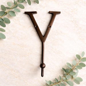 Könnte beinhalten: Ein bronzefarbener Metall-Wandhaken in Form des Buchstabens "V" mit einem Haken am unteren Ende und einem kleinen Kugeldetail. Der Hintergrund ist eine helle, strukturierte Oberfläche mit grünem Laub.