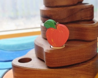 May include: A wooden stacking toy with a red apple on top. The toy is made of natural wood and has a smooth finish. The apple is painted red with a green leaf.