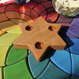 Puede incluir: Un portavelas de madera en forma de estrella con tres aberturas circulares, apoyado sobre una base de madera colorida con un patrón de arcoíris. La estrella es de color marrón claro y la base presenta tonos de rojo, naranja, amarillo, verde, azul y morado.