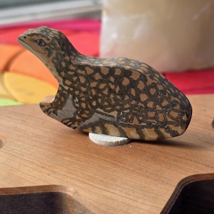 以下が含まれることがあります： 茶色と黒の斑点模様が施された、手描きの小さな木製のトカゲの置物。トカゲは明るい茶色の星型の木製台座の上に置かれており、詳細な目と質感のある鱗のような外観が特徴です。