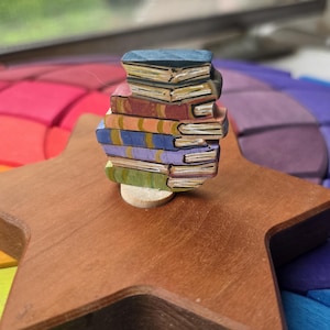 May include: A small, hand-painted wooden figurine of a stack of six books in various colors including blue, red, yellow, purple, and green, resting on a brown wooden star-shaped base with a light-colored circular support underneath the books.