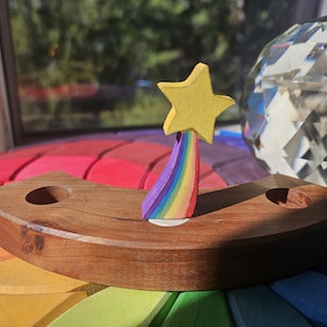 May include: Wooden rainbow-themed toy with a yellow star and a rainbow arc. The toy is set on a colorful, circular base. A clear, faceted glass object is in the background. The wooden base has two holes.