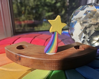 May include: Wooden rainbow-themed toy with a yellow star and a rainbow arc. The toy is set on a colorful, circular base. A clear, faceted glass object is in the background. The wooden base has two holes.
