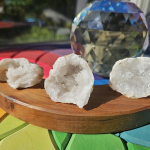 May include: Three white quartz geodes sit on a curved wooden tray. The geodes have a crystalline structure and are displayed against a backdrop of colorful wooden blocks and a faceted glass sphere.