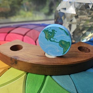 May include: A wooden toy depicting a blue and green globe, placed on a curved, brown wooden base with two holes. The base sits on a colourful, rainbow-themed wooden block set. A faceted crystal sphere is in the background.