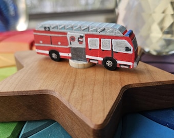 May include: A miniature red and white fire truck toy with a silver roof, sitting on a wooden star-shaped base. The toy has black wheels and is painted with details. The base is placed on a colorful, rainbow-themed surface.