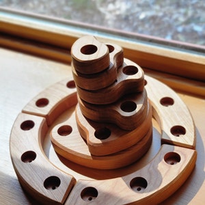Puede incluir: Un juguete de apilar de madera con una base circular y varios anillos que encajan entre sí. Los anillos tienen forma de círculos irregulares con agujeros. El juguete está hecho de madera natural y tiene un color marrón claro.