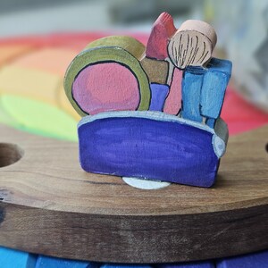 May include: A wooden toy depicting a makeup kit, with a pink blush, a green and pink compact, and a blue container. The toy is set on a wooden base with a rainbow background.