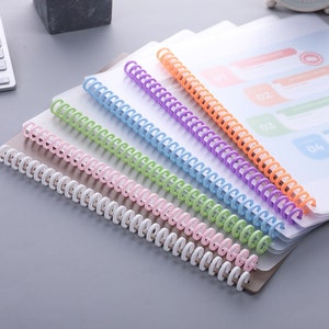 May include: A collection of colorful plastic coil binding spines in white, pink, green, blue, purple, and orange. The spines are arranged diagonally across a stack of clear document covers, suggesting an office or school supply theme.