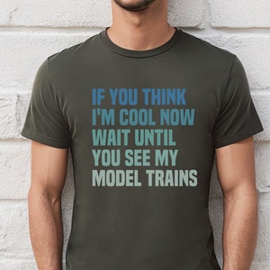 May include: A dark green t-shirt with the text "If you think I'm cool now wait until you see my model trains" printed in blue and green.