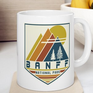 May include: White ceramic mug with a retro-style graphic of mountains, trees, and a sun. The text "BANFF NATIONAL PARK" is printed in blue on the mug.