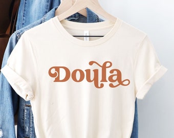 Könnte beinhalten: Ein weißes T-Shirt mit dem Wort "Doula" in retro-orangefarbener Schrift. Das Shirt hängt an einem Kleiderbügel und hat eine natürliche Farbe.