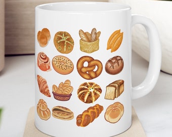 Könnte beinhalten: Weiße Keramiktasse mit einer bunten Illustration verschiedener Brote und Gebäckstücke. Die Tasse hat einen Henkel auf der rechten Seite.