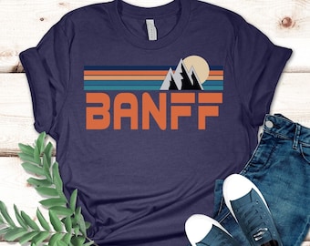 May include: A heather navy blue t-shirt with a retro graphic design featuring mountains, a sun, and the word "BANFF" in orange.