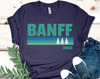 May include: A navy blue t-shirt with a white graphic of the word "BANFF" in green letters with a green and blue striped design and three white pine trees. The text "CANADA" is below the trees.