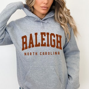 Pode incluir: Uma sudadera com capuz cinza mescla com uma grande estampa marrom que diz "RALEIGH NORTH CAROLINA".