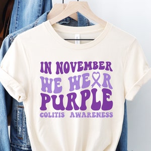 May include: A white t-shirt with the text "In November We Wear Purple" in a retro font. The text is in purple and includes a purple ribbon symbol. The text "Colitis Awareness" is below the main text.