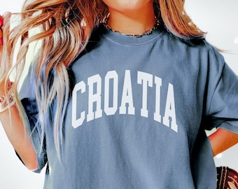 Op de afbeelding: Een blauw t-shirt met het woord "CROATIA" in witte blokletters. Het shirt is effen van kleur en heeft korte mouwen. Het model draagt een gouden ketting en armbanden. Bruine cowboylaarzen zijn zichtbaar op de achtergrond.