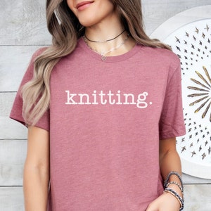 May include: A mauve coloured t-shirt with the word "knitting." printed in white on the front.