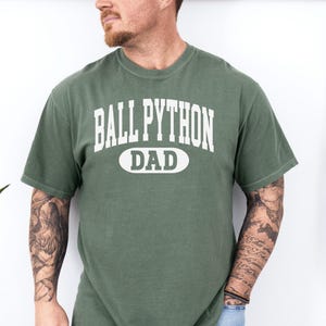Könnte beinhalten: Grünes T-Shirt mit dem Text "BALL PYTHON DAD" in weißen Blockbuchstaben. Das Wort "DAD" befindet sich in einem weißen Oval. Das Shirt ist einfarbig grün und hat kurze Ärmel.