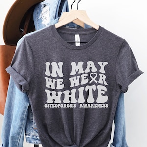 May include: Dark heather grey t-shirt with the text "In May We Wear White" in a retro font. The text is in white and includes a white ribbon symbol. Below the text is "Osteoporosis Awareness".