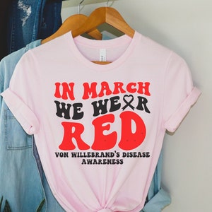May include: Pink t-shirt with the text "In March We Wear Red" in red and black letters. The text "Von Willebrand's Disease Awareness" is printed below in black letters.