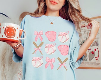 May include: A light blue t-shirt with a graphic design of pink bows, baseball bats, baseballs, and baseball gloves.