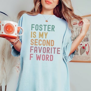 Pode incluir: Uma camiseta azul claro com o texto "Foster is my second favorite F word" em cores do arco-íris.