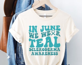 May include: A cream-colored t-shirt with the text "In June We Wear Teal Scleroderma Awareness" in teal blue lettering. The text is in a retro font style.