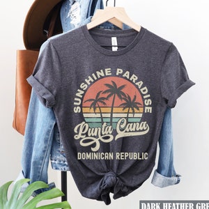 May include: Dark heather grey t-shirt with a vintage-style graphic of palm trees and a sunset. The graphic says "Sunshine Paradise Punta Cana Dominican Republic".