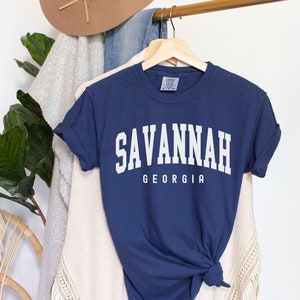 Op de afbeelding: Marineblauw T-shirt met het woord "SAVANNAH" in witte blokletters boven "GEORGIA". Het shirt hangt aan een houten hanger met andere kleding en een hoed op de achtergrond.