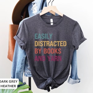 May include: Dark grey heather t-shirt with the text "Easily Distracted By Books And Yarn" in a retro rainbow color scheme.