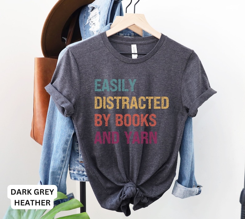 May include: Dark grey heather t-shirt with the text "Easily Distracted By Books And Yarn" in a retro rainbow color scheme.