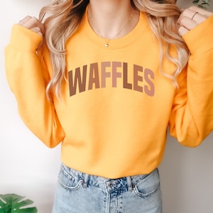 May include: A yellow sweatshirt with the word "WAFFLES" printed in brown, orange, and pink letters.